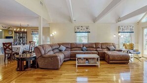 A spacious living room with a large sectional sofa, wooden flooring, and a dining area with a table and chairs. - Wonderful waterfront getaway with furnished deck, pond access, kayaks & WiFi (Cotuit)