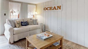 A living room with a beige sofa, a coffee table, and a wall-mounted sign that reads 'OCEAN'. - Classic Cape Cod home with Patio and Firepit - Walk to the Beach (Dennis Port)