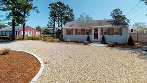 A single-story house with a front porch, a driveway, and a well-maintained lawn. - Classic Cape Cod home with Patio and Firepit - Walk to the Beach (Dennis Port)