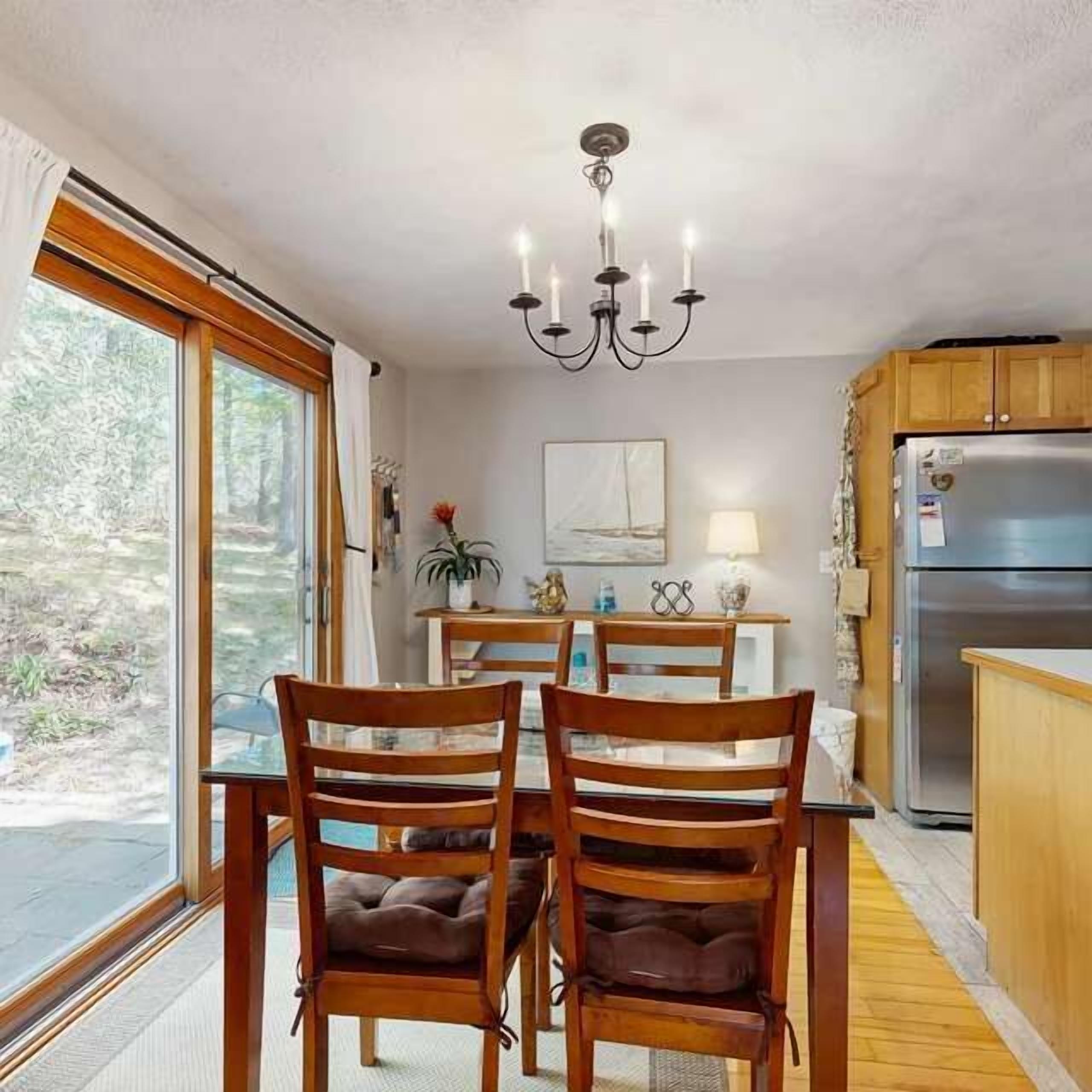 Un comedor con mesa, sillas, una araña, refrigerador y ventana con cortinas.