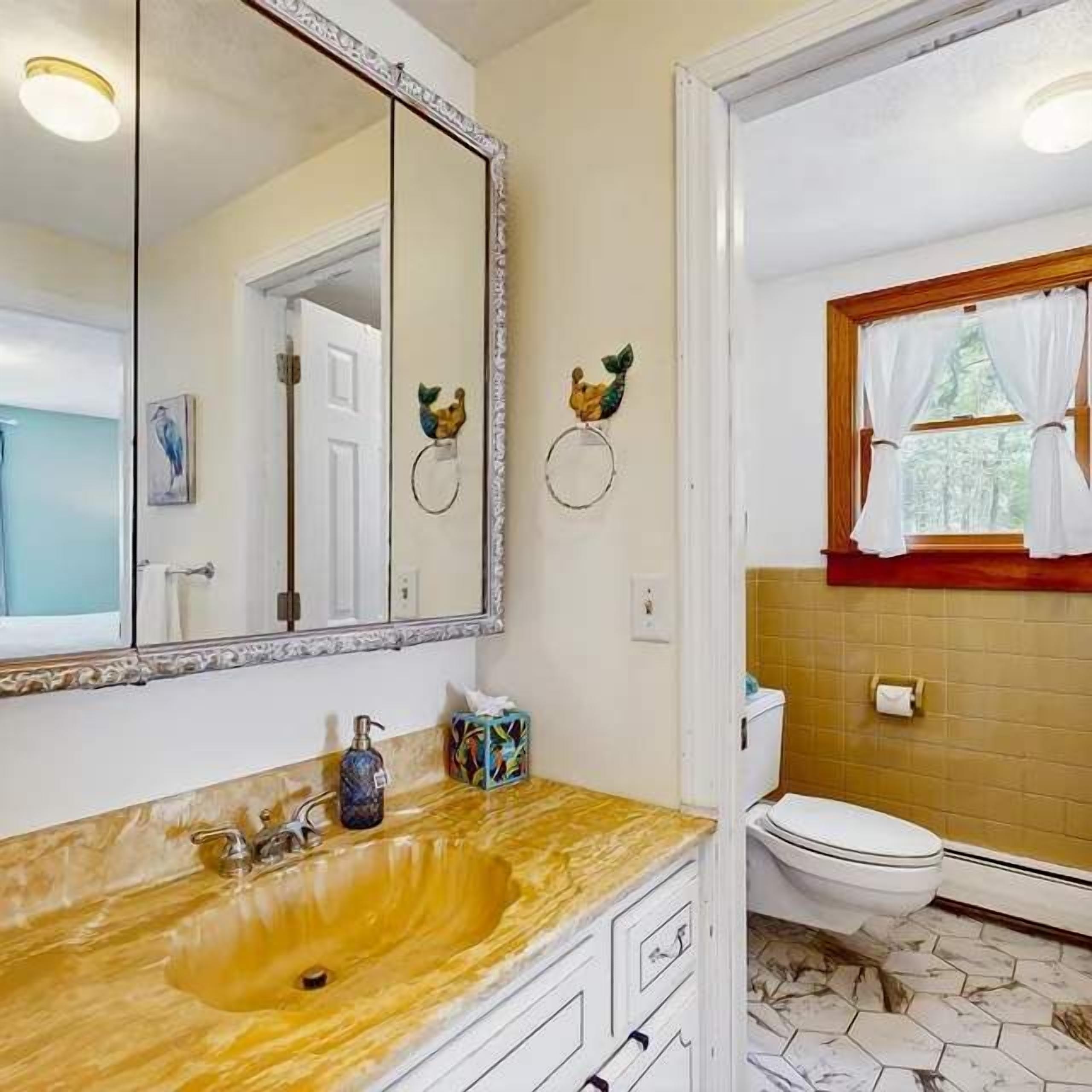 Un baño con lavamanos de mármol, un espejo grande, inodoro y ventana con cortinas.