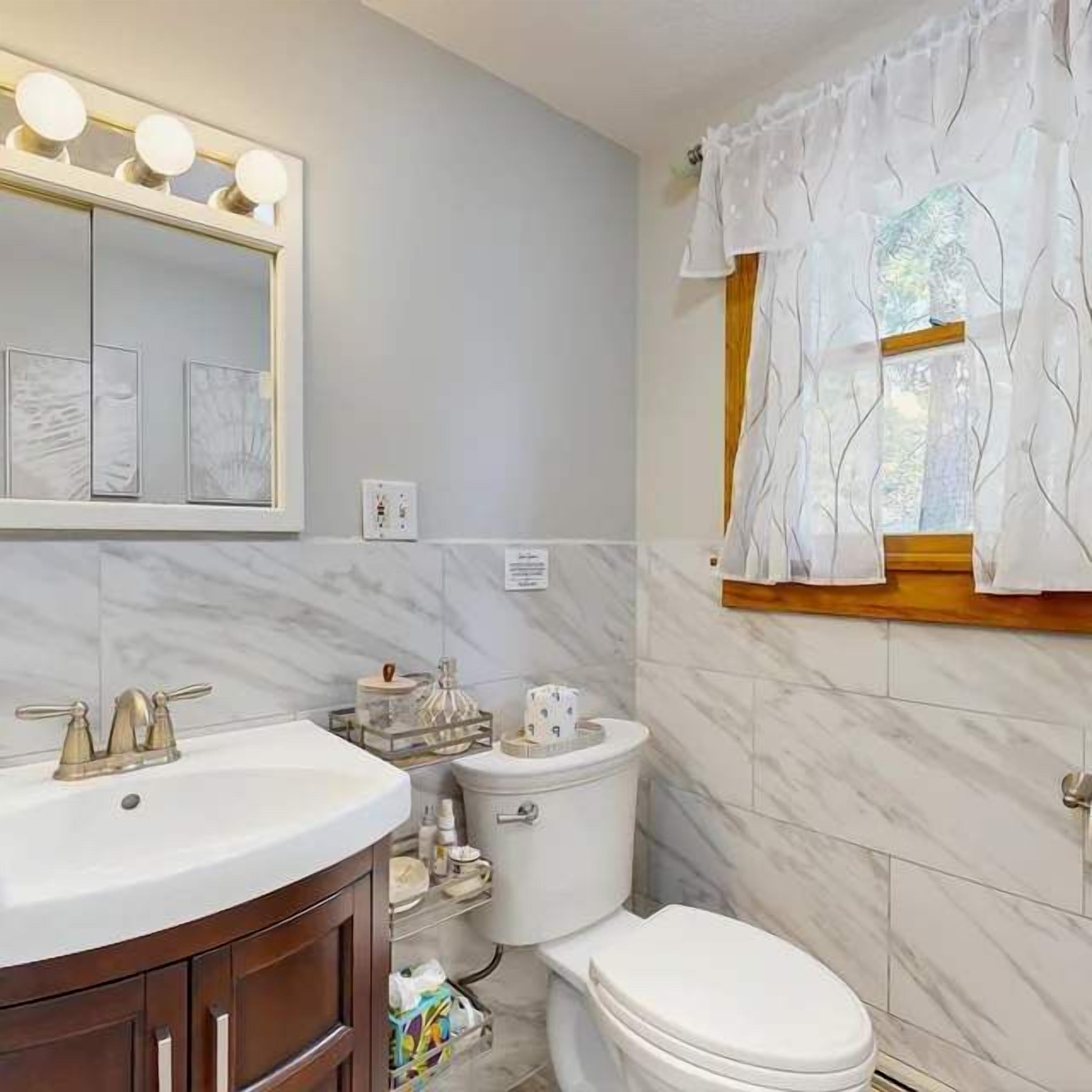 Un baño con tocador, lavabo, espejo con luces, inodoro y ventana con cortinas.