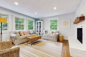 A living room with two sofas, a coffee table, a bookshelf, and a fireplace. - Beautiful bright home with firepit, deck & backyard - walk to beach (Falmouth)