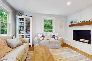 A living room with a sofa, a coffee table, a bookshelf, and a fireplace. - Beautiful bright home with firepit, deck & backyard - walk to beach (Falmouth)