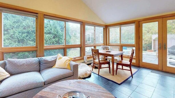 A spacious room with a large window, a sofa, a dining table with chairs, and a wooden coffee table. - Quaint home w/ large backyard, outdoor shower & private grill near Katama Bay (Edgartown)
