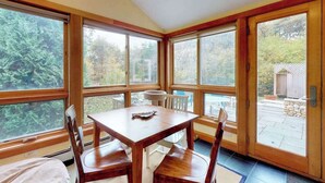 A sunlit room with a wooden table and chairs, large windows, and a view of trees. - Quaint home w/ large backyard, outdoor shower & private grill near Katama Bay (Edgartown)