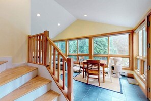 A staircase with wooden railings leading to a room with large windows, a dining table, and a cat tree. - Quaint home w/ large backyard, outdoor shower & private grill near Katama Bay (Edgartown)
