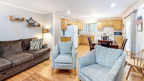 Ein Wohnzimmer mit einer Couch, zwei Sesseln, einem Couchtisch und einem Essbereich mit Tisch und Stühlen. Im Hintergrund befindet sich eine Küche mit Holzschränken und einem Kühlschrank.