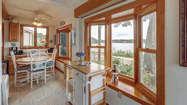 A kitchen with wooden cabinets, a dining table with chairs, a window with a view of the water, and a ceiling fan. - Family Friendly Falmouth Beach House with Ocean Views and BBQ Grill (Falmouth)