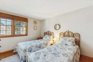 A bedroom with two beds, a window with blinds, a bedside table with a lamp, and a wall-mounted mirror.