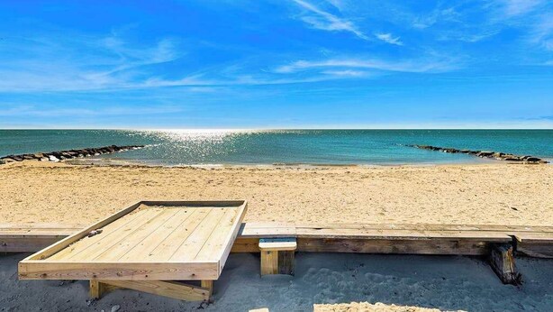 En træterrasse med bænk og bord, med udsigt over en strand med klart, blåt vand og en stenmolle.