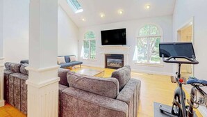 A spacious living room with a fireplace, a television, and a stationary bike. - Inviting home with charming outdoor area & firepit - short drive downtown (East Falmouth)