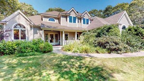 A two-story house with a front porch, a covered entryway, and a well-maintained lawn. - Inviting home with charming outdoor area & firepit - short drive downtown (East Falmouth)