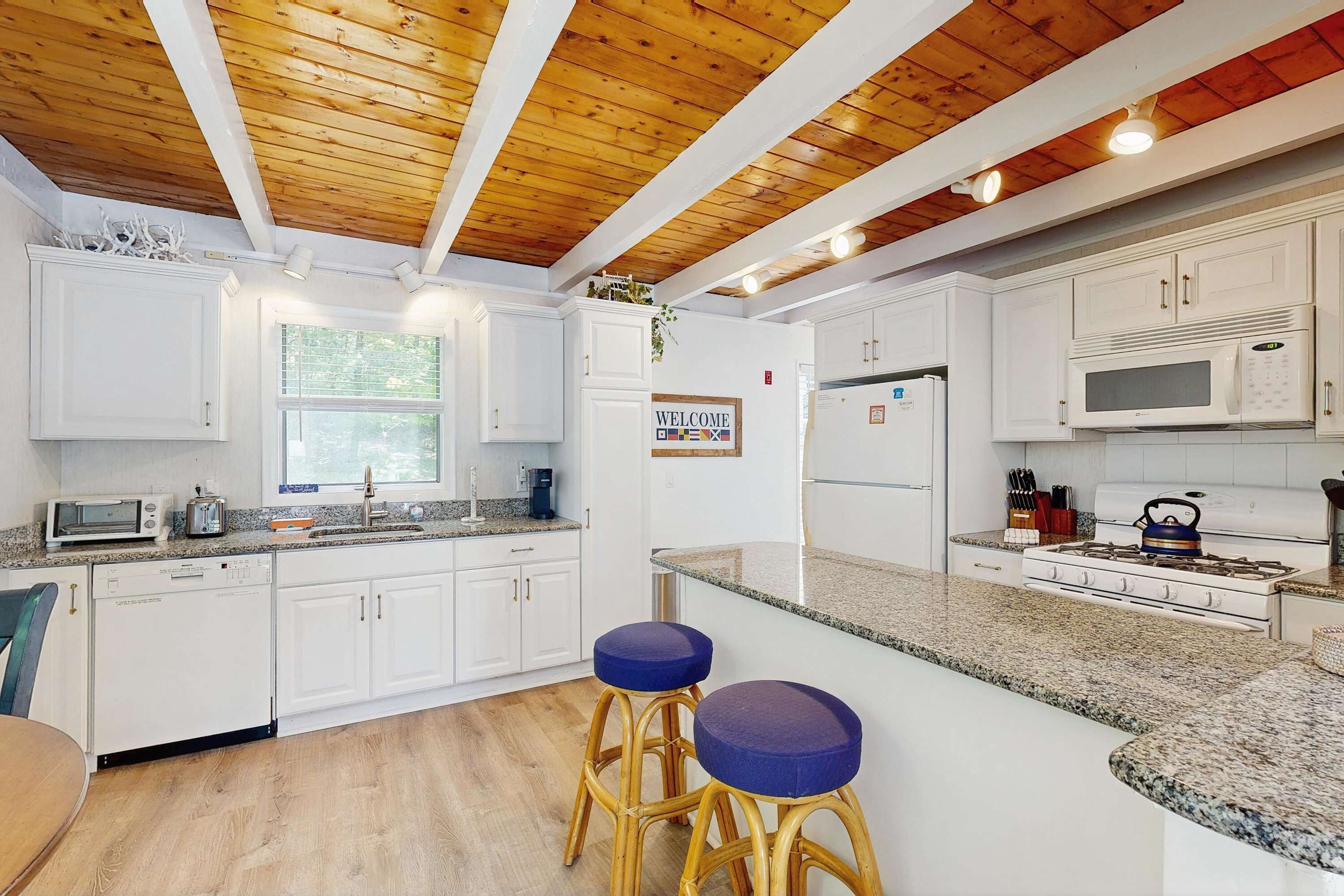 Una cocina moderna con armarios blancos, microondas integrado, estufa y lavaplatos. Hay dos taburetes azules y una ventana con persianas.