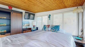 Un dormitorio con techo de madera, una cama con ropa de cama blanca, un televisor montado en la pared y una ventana con persianas.