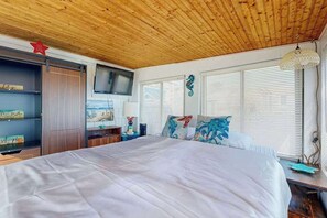 A bedroom with a wooden ceiling, a bed with white bedding, a TV mounted on the wall, and a window with blinds. - Charming cottage with private beach, patio, & full kitchen - near golf & park (Dennis)