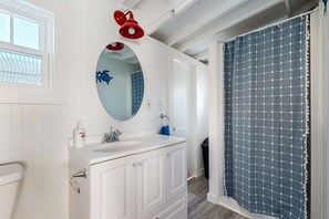 A bathroom with a white vanity, a round mirror, a red showerhead, and a blue shower curtain. - Charming cottage with private beach, patio, & full kitchen - near golf & park (Dennis)