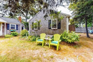 A house with a sloped roof, a front porch with two Adirondack chairs, and a small table. - Cape-style cottage in a small village community w/ an outdoor shower (Harwich Port)