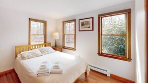 Un dormitorio con cama de madera, dos ventanas, un cuadro en la pared y una mesita de noche con lámpara.