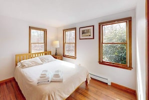 A bedroom with a wooden bed, two windows, a wall-mounted picture, and a nightstand with a lamp.