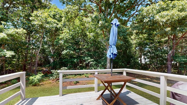A wooden deck with a table and an umbrella, surrounded by trees. - Cape Getaway -walk to restaurants & shops, near bay & ocean beaches & rail trail (Eastham)