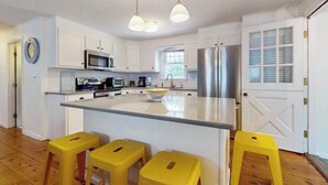 A modern kitchen with white cabinetry, a central island, stainless steel appliances, and yellow stools. - Cape Getaway -walk to restaurants & shops, near bay & ocean beaches & rail trail (Eastham)