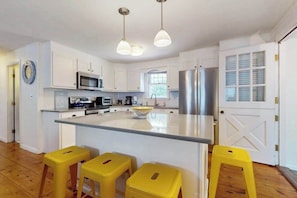 A modern kitchen with white cabinetry, a central island, stainless steel appliances, and yellow stools. - Cape Getaway -walk to restaurants & shops, near bay & ocean beaches & rail trail (Eastham)