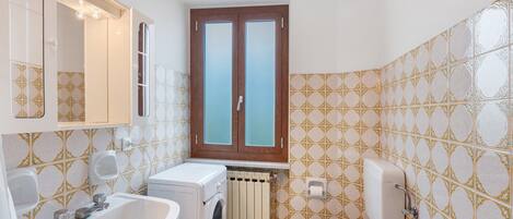A compact bathroom with a sink, washing machine, toilet, and a window with a view.
