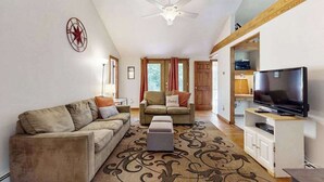 Una sala de estar con sofá, sillón, reposapiés, televisor, ventilador de techo y alfombra estampada.
