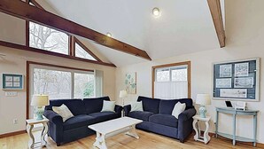 A living room with two dark sofas, a white coffee table, and a small side table with a lamp. There are large windows and a ceiling with exposed beams.