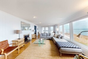A spacious living room with a large window, a sofa, a coffee table, and a fireplace. - Waterfront Hyannis Haven – Walk to Kalmus Beach (Hyannis)