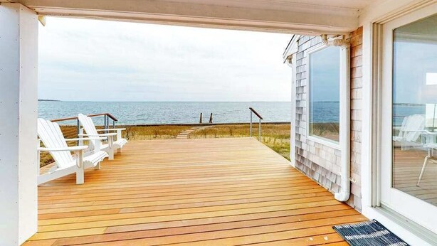 A wooden deck with white chairs, a view of the sea, and a glass door leading to the interior. - Waterfront Hyannis Haven – Walk to Kalmus Beach (Hyannis)