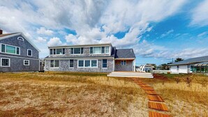 A two-story house with a stone facade, a wooden deck leading to the front door, and a white picket fence. - Waterfront Hyannis Haven – Walk to Kalmus Beach (Hyannis)