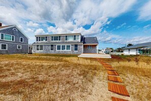 A two-story house with a stone facade, a wooden deck leading to the front door, and a white picket fence. - Waterfront Hyannis Haven – Walk to Kalmus Beach (Hyannis)
