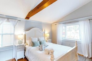 Ein Schlafzimmer mit Holzboden, einem weißen Bett mit vielen Kissen, Nachttischlampen und einem Fenster mit Vorhängen.