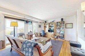 A spacious living room with a sofa, chairs, a coffee table, a bookshelf, and a television. - Spacious home on golf course with 4 outdoor spaces overlooking the Sound (Mashpee)