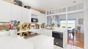 A modern kitchen with white cabinetry, a dishwasher, and a dining area with a table and chairs. - Spacious home on golf course with 4 outdoor spaces overlooking the Sound (Mashpee)