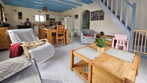 Een ruime woonruimte met een houten salontafel, een grijze fauteuil en een eettafel met stoelen. Een trap met blauwe leuningen leidt naar een hoger niveau.