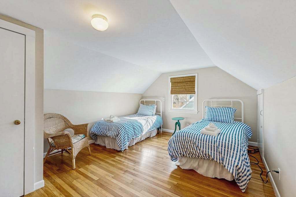 A bedroom with two beds, a wicker chair, a small table, and a window with a blind.