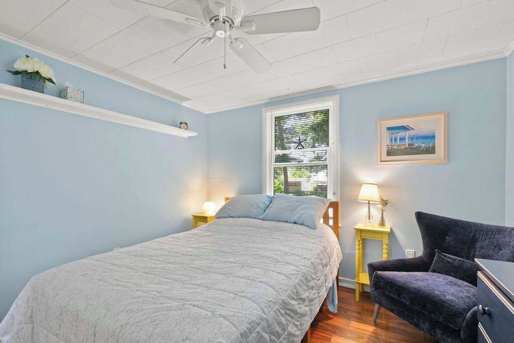 Une chambre à coucher comprenant un lit, une chaise, une table de chevet, une lampe, un ventilateur de plafond, une fenêtre et une étagère avec des objets décoratifs.