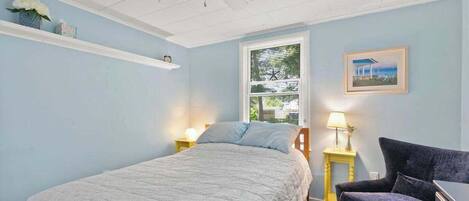 Une chambre à coucher comprenant un lit, une chaise, une table de chevet, une lampe, un ventilateur de plafond, une fenêtre et une étagère avec des objets décoratifs.