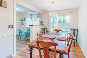 A dining area with a wooden table and chairs, a chandelier, and a view of the backyard through a sliding glass door. - Private house close to beach & playground with grill, outdoor shower, & patio (Falmouth)