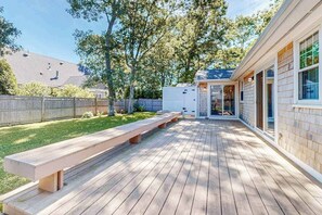 A wooden deck with benches, a covered area, and a view of a house with a fenced backyard. - Private house close to beach & playground with grill, outdoor shower, & patio (Falmouth)