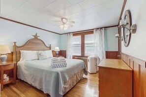 A bedroom with a bed, a ceiling fan, a radiator, a dresser, and a window with curtains.