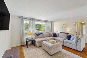A modern living room with a sectional sofa, a small table, and a TV mounted on the wall. - Neighborhood charmer with deck, W/D, Outdoor shower, Dog friendly, Wifi (Falmouth)