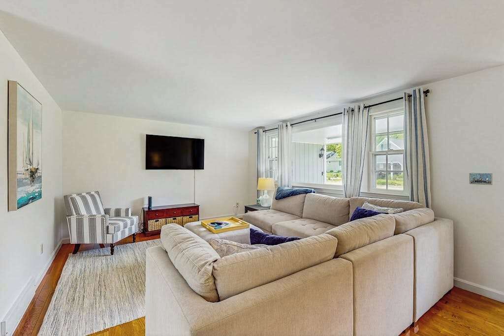 A living room with a sectional sofa, a flat-screen TV, a striped armchair, a small table with a lamp, and a window with curtains.