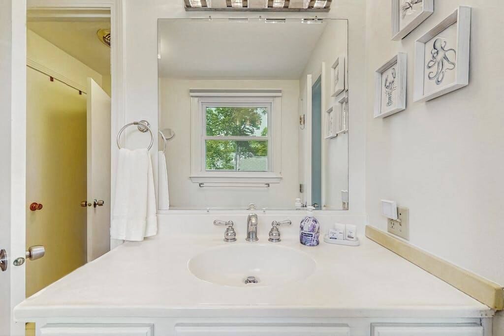 Une salle de bain avec un lavabo blanc, deux robinets, un miroir et des œuvres d’art encadrées accrochées au mur.