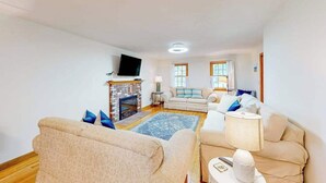 A living room with a fireplace, sofas, a TV, and a window. - Our House in West Falmouth | House in Falmouth with private beach access (Falmouth)