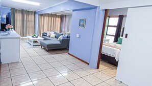Una sala moderna con sofá, una mesa de centro y televisión. Se observa una zona de cocina con mostrador y fregadero. El piso es de baldosas y las paredes son de color azul claro.