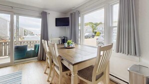Uma sala de jantar com mesa de madeira, cadeiras, televisão de tela plana e janelas amplas com cortinas.